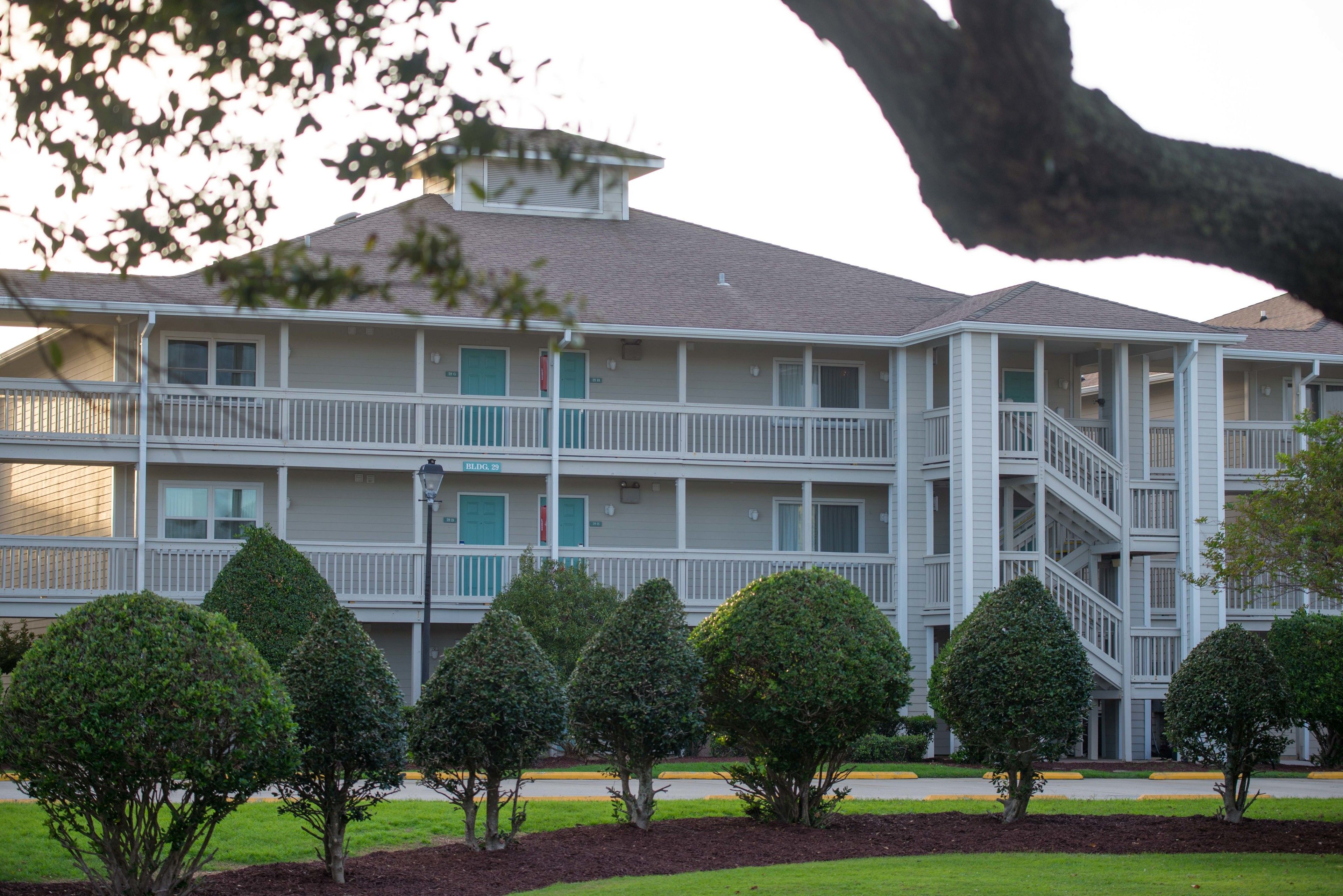 A multi-story hotel with a balcony on the second floor, a covered outdoor area, and a well-maintained lawn with trimmed bushes.