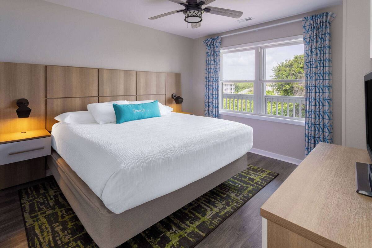 A neatly made bed with a white comforter and a teal pillow, a wooden headboard, a bedside table with a lamp, a window with blue curtains, and a ceiling fan.