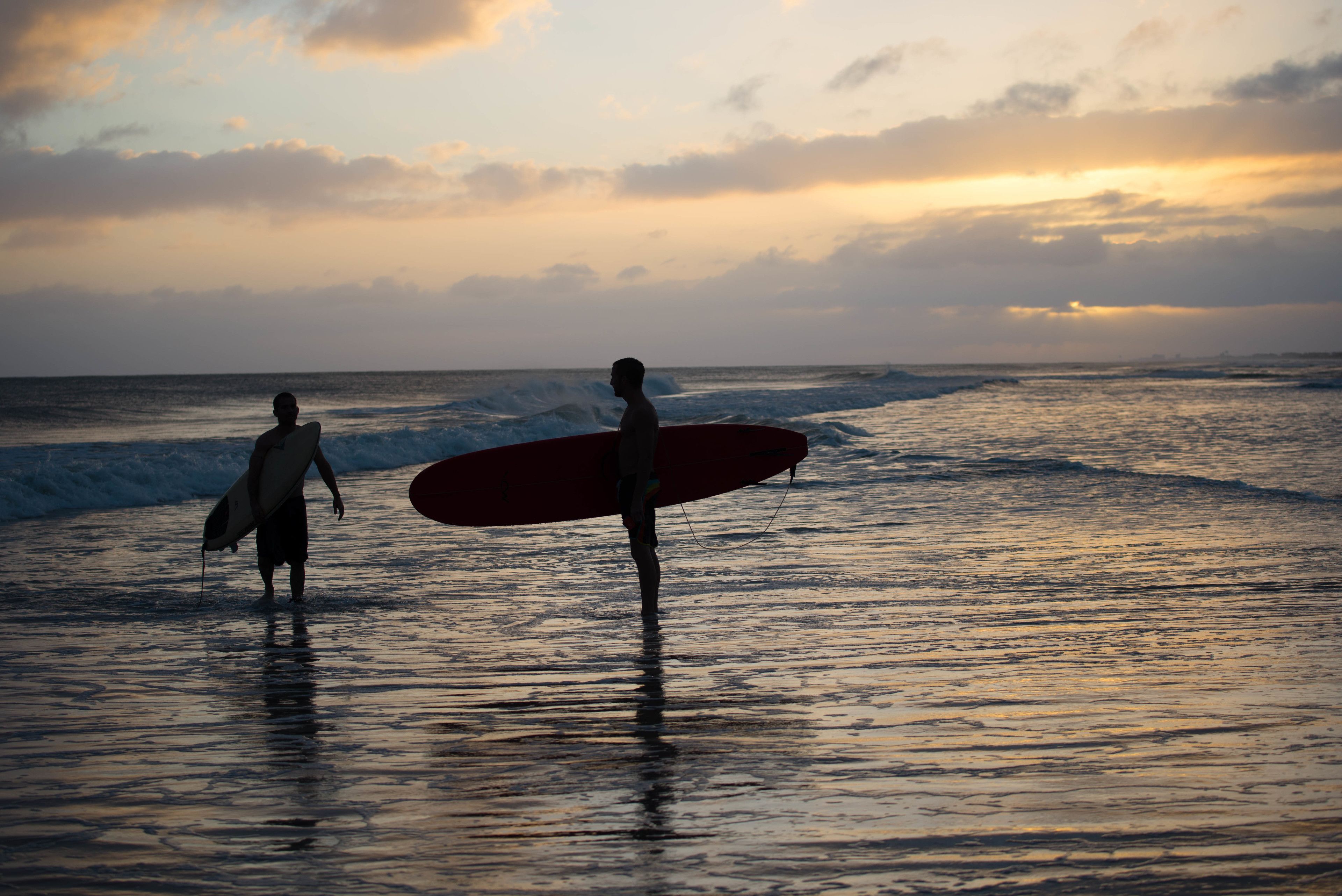 Two people walking on a beach with surfboards, under a sunset sky.