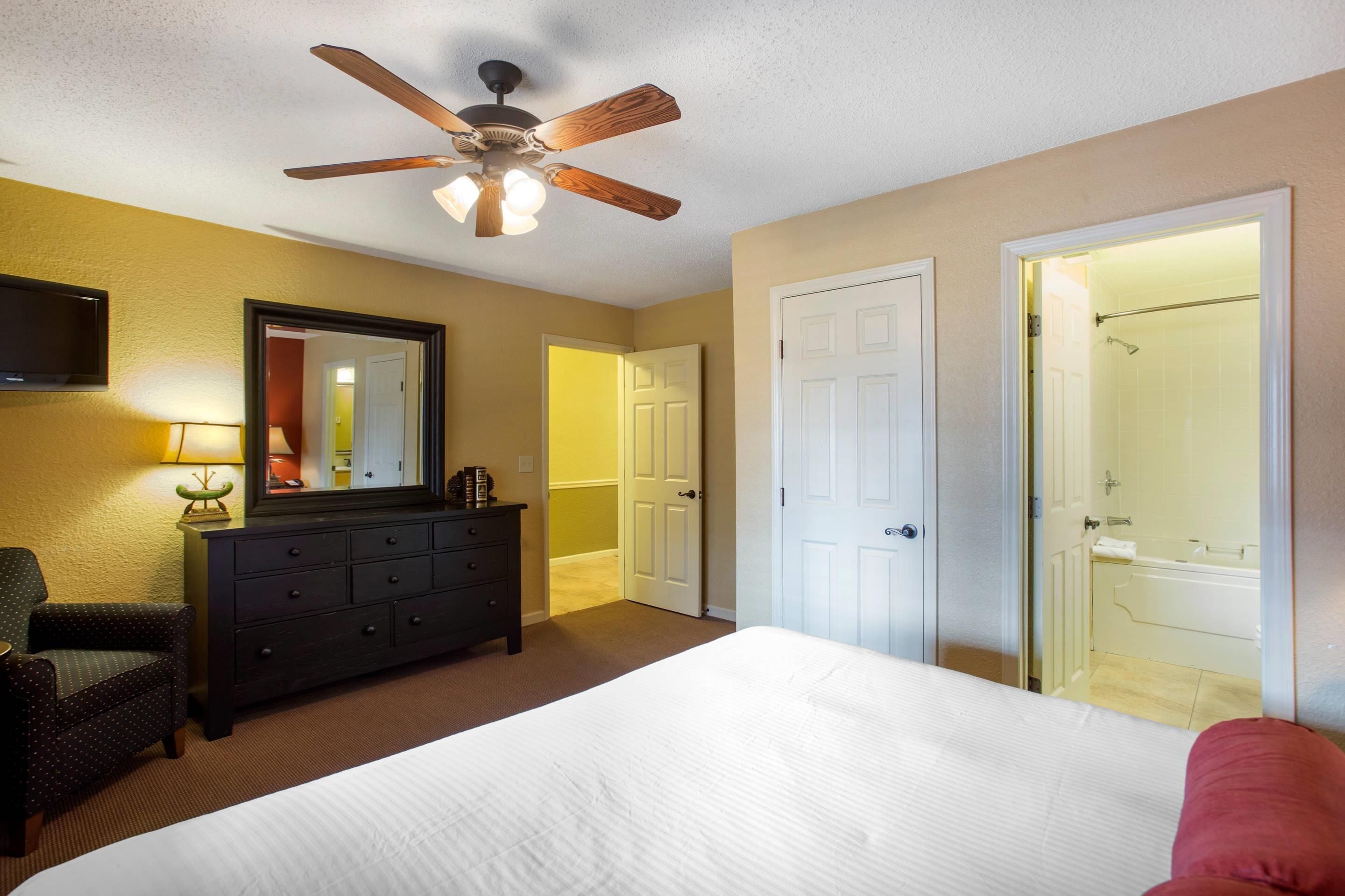 A hotel room with a bed, a ceiling fan, a dresser, a mirror, and two doors leading to a bathroom.