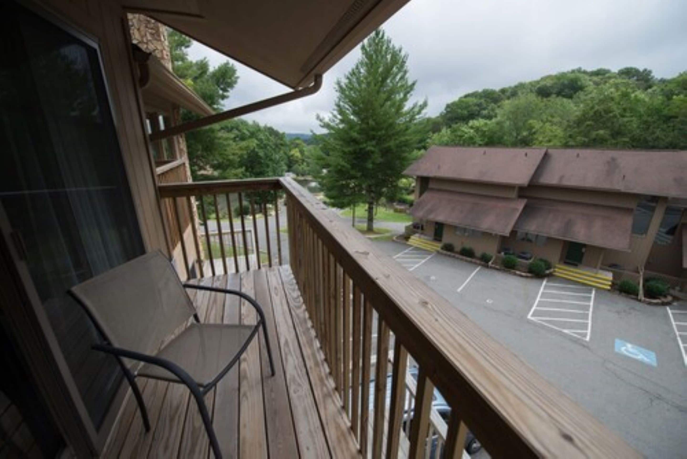 A wooden deck with a chair, overlooking a parking lot and a building with a brown roof.