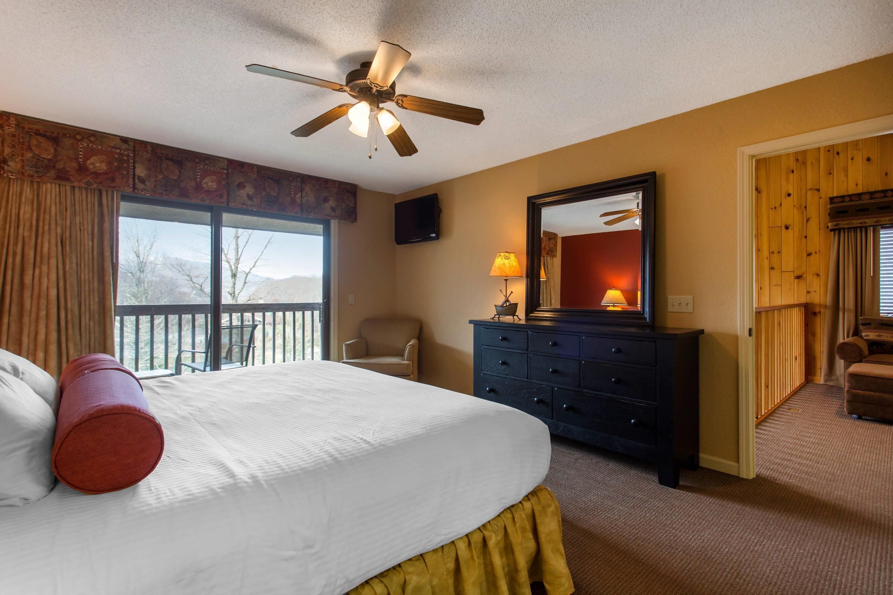 A hotel room with a bed, a ceiling fan, a dresser, a chair, a television, and a view of the outdoors.