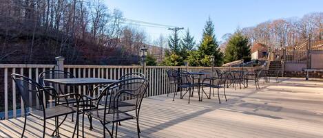 An outdoor deck with metal tables and chairs, surrounded by a wooden railing and trees.
