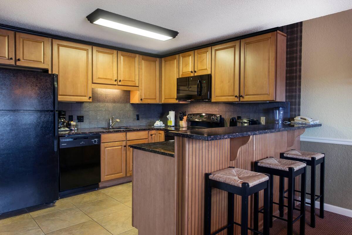 A kitchen with wooden cabinets, a black refrigerator, a microwave, a coffee maker, and a bar area with stools.