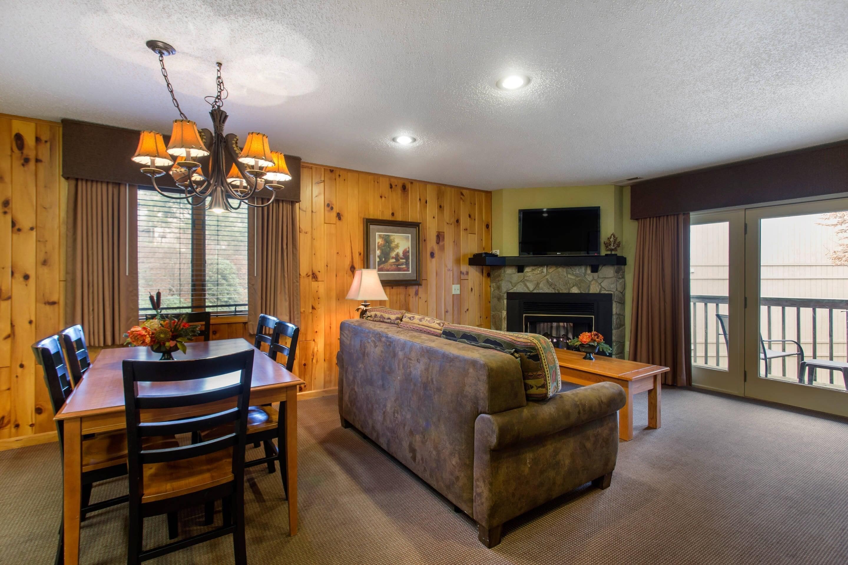 A cozy living room with a fireplace, wooden walls, a dining area with a chandelier, and a balcony with outdoor seating.