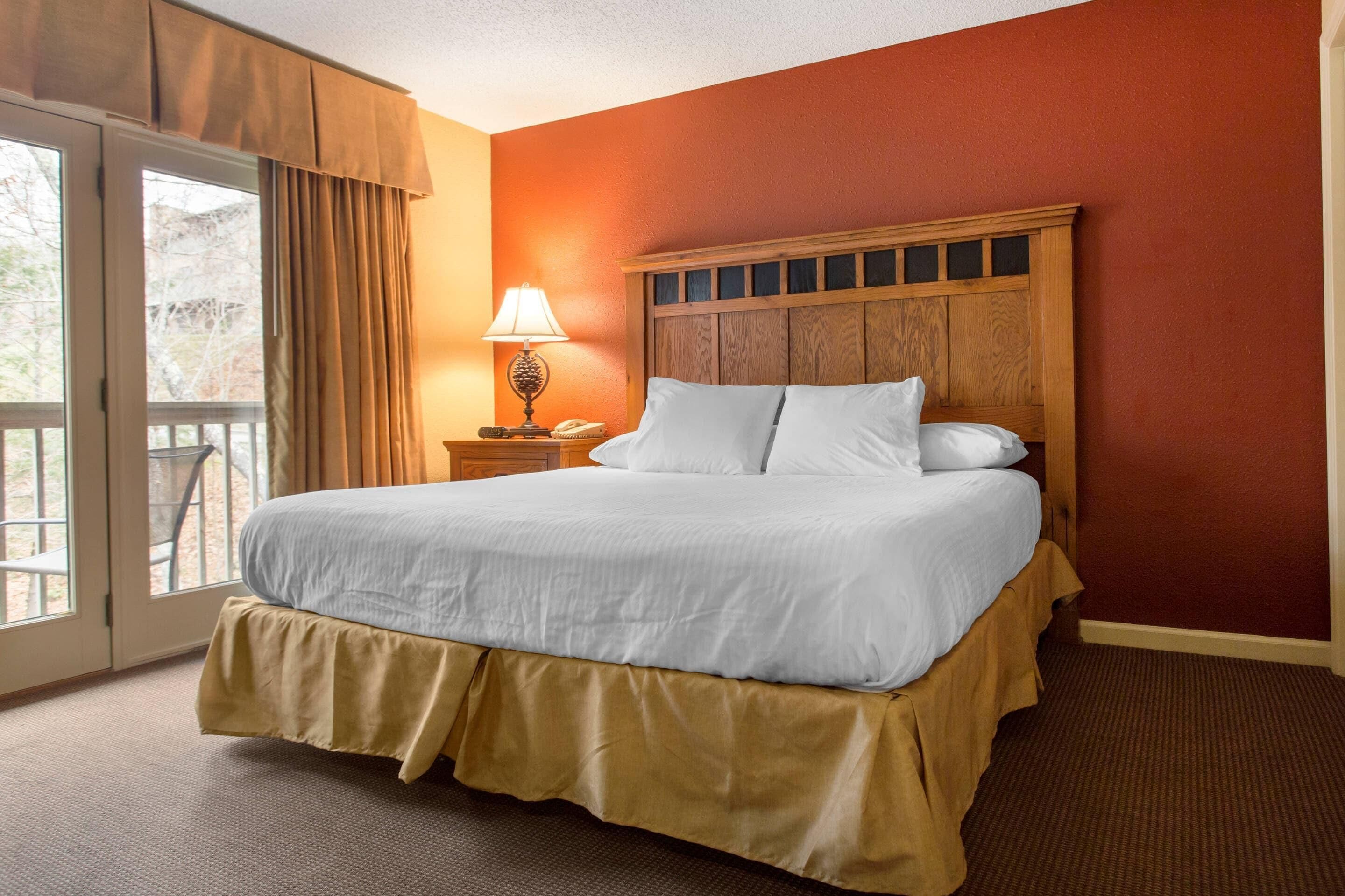 Ein Hotelzimmer mit einem großen Bett, einem hölzernen Kopfteil, einem Nachttisch mit Lampe und Blick durch eine Glastür.