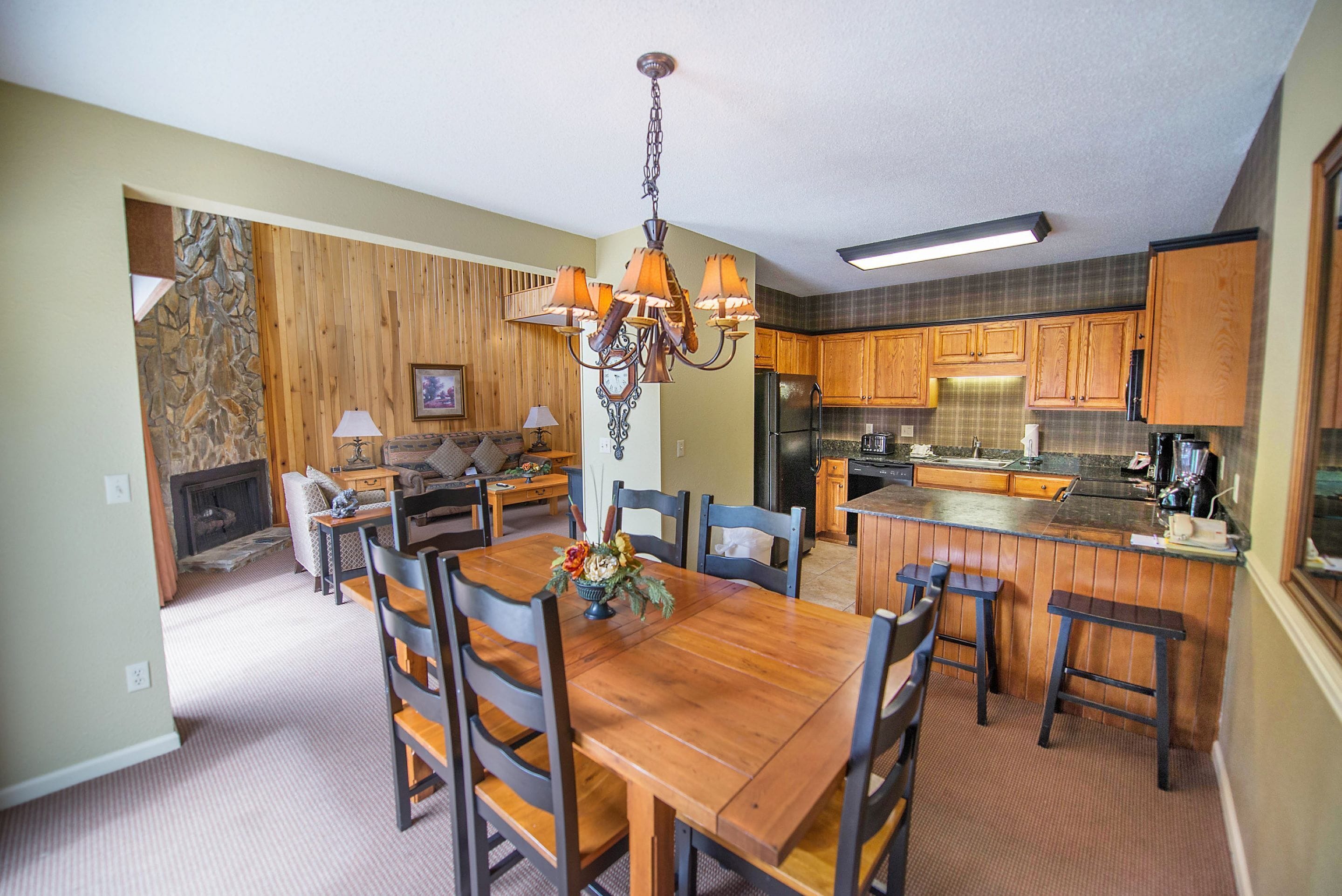 A spacious dining area with a wooden table and chairs, a chandelier, and a kitchen with wooden cabinets and a refrigerator.
