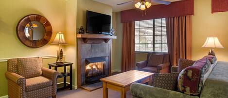 Una sala de estar con chimenea, televisor, ventilador de techo y un gran ventanal con cortinas.