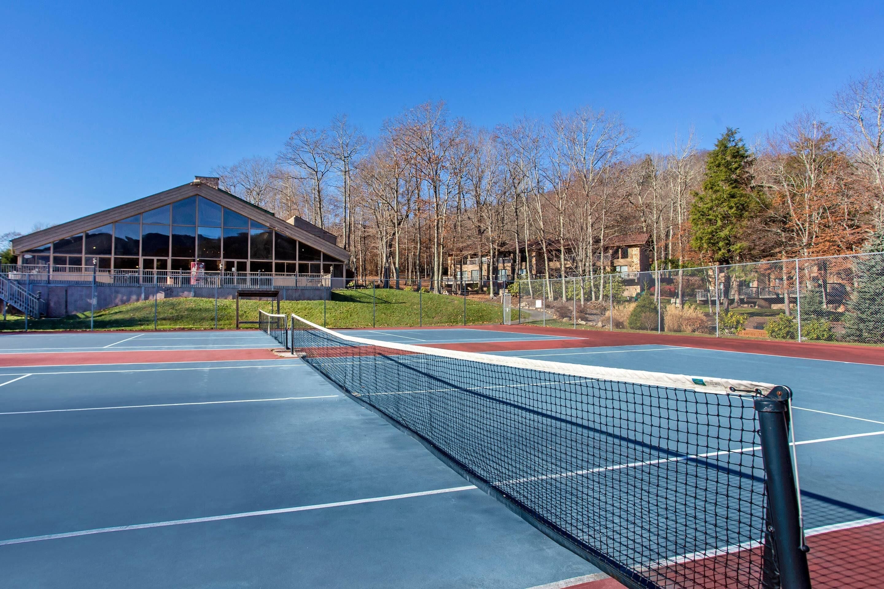 An outdoor tennis court with a net, surrounded by a fence, and a building with large windows in the background.