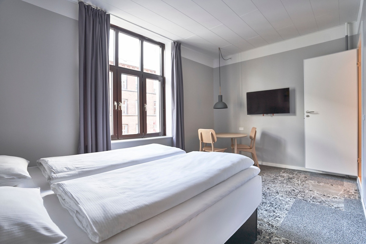 A hotel room with two beds, a desk, a chair, a TV, and a window with curtains.