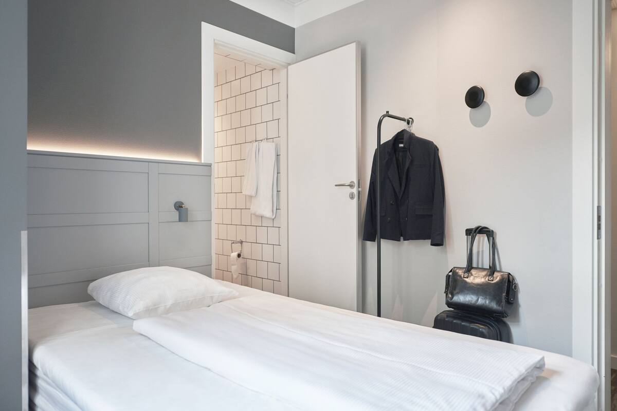 Una camera d'albergo moderna con un letto rifatto, uno spazio doccia e una valigia su un carrello.