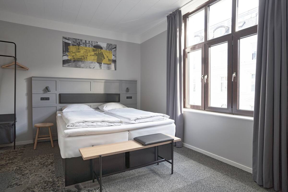 Una camera d'albergo con un letto, una scrivania, una sedia, un attaccapanni e una finestra con tende.