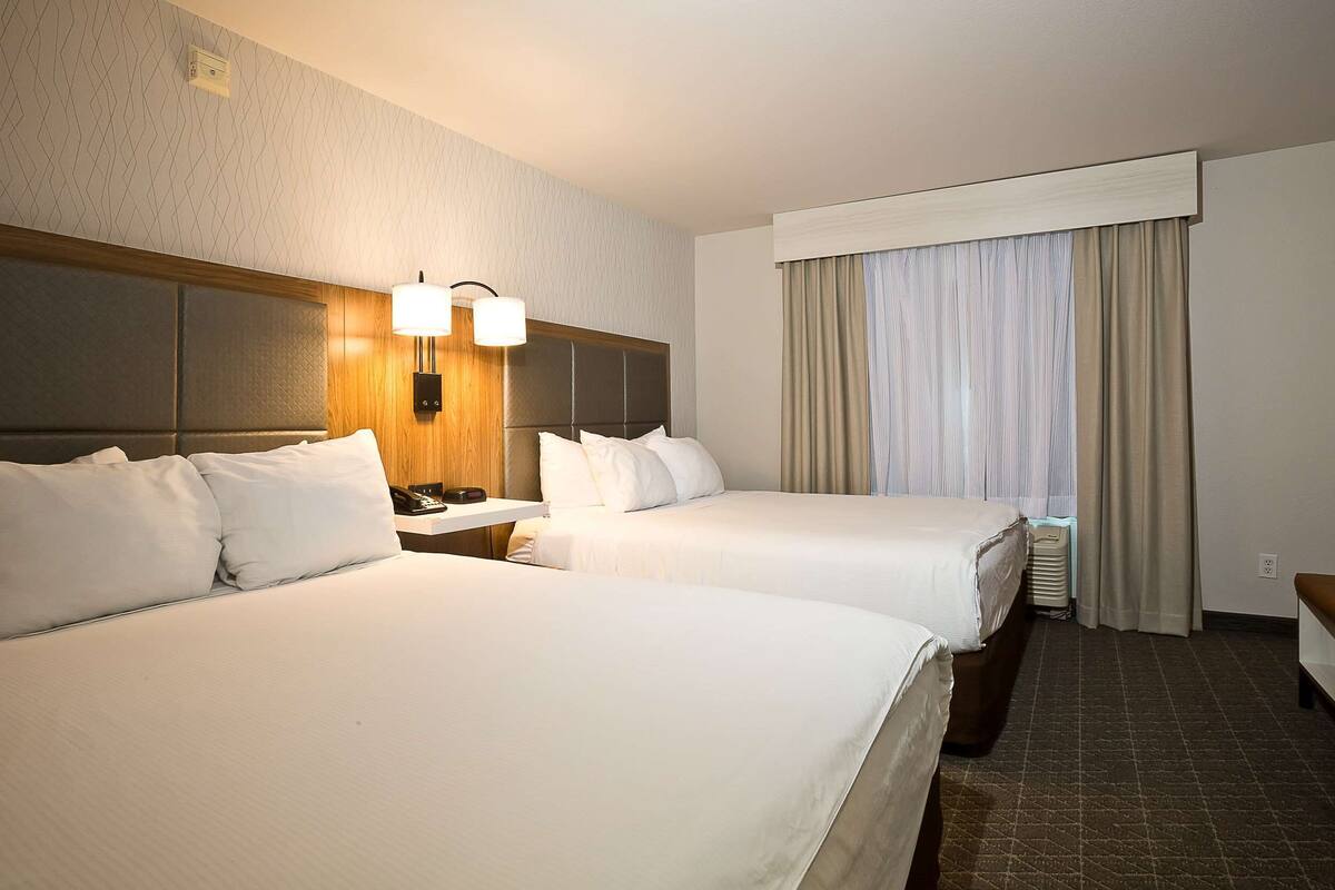 A hotel room with two beds, a nightstand with a lamp, a window with curtains, and a wall with a textured pattern.