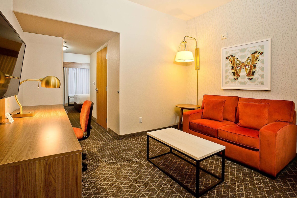 A hotel room with a wooden desk, an orange sofa, a white coffee table, a floor lamp, and a butterfly artwork on the wall.