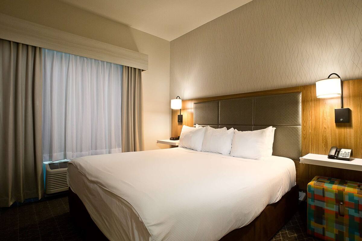 A hotel room with a large bed, two bedside tables with lamps, a small table with a phone, and a colorful ottoman.