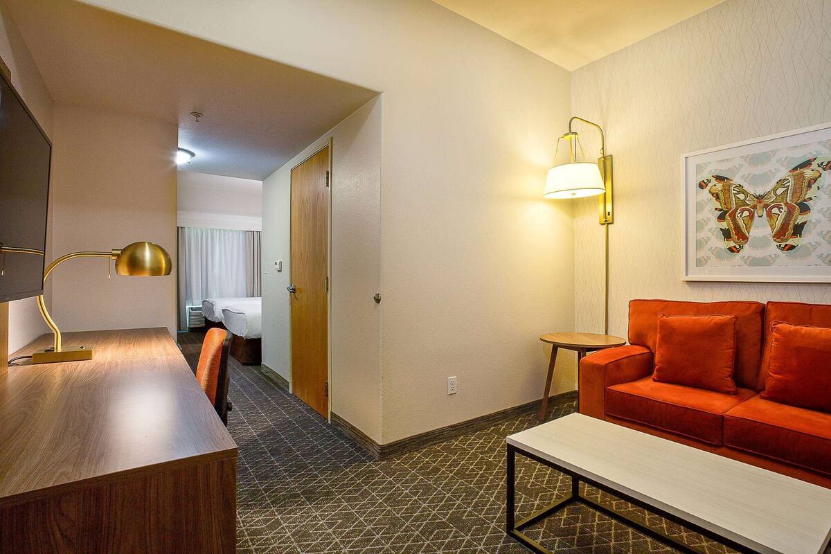 A hotel room with a wooden desk, a red sofa, a white coffee table, a wall-mounted lamp, and a framed butterfly artwork.