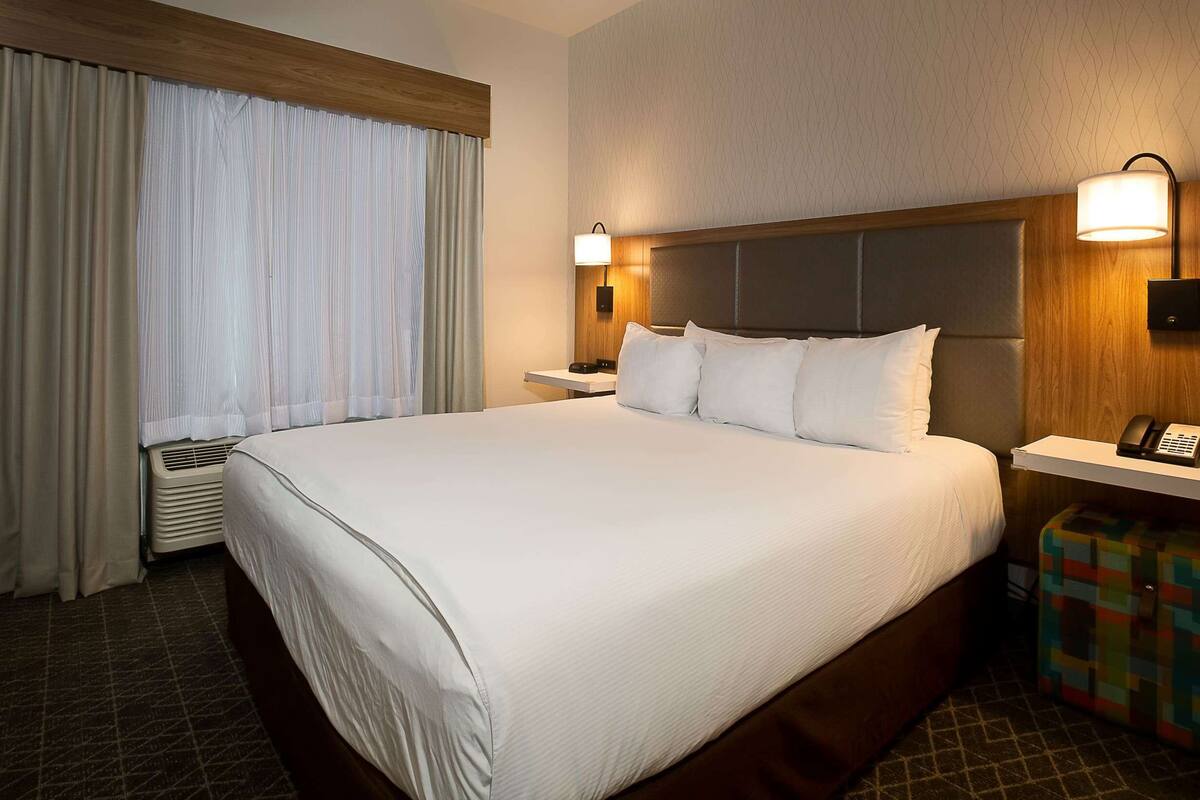 A hotel room with a large bed, a bedside table with a lamp, a telephone, and a colorful suitcase.