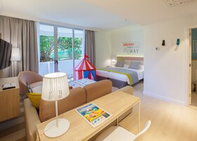 Habitación de hotel con cama, escritorio, silla, lámpara, ventana con cortinas y una pared con una frase motivacional.
