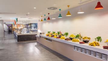 Zona de bufé con diversas frutas y verduras, una larga barra y lámparas colgantes de colores.