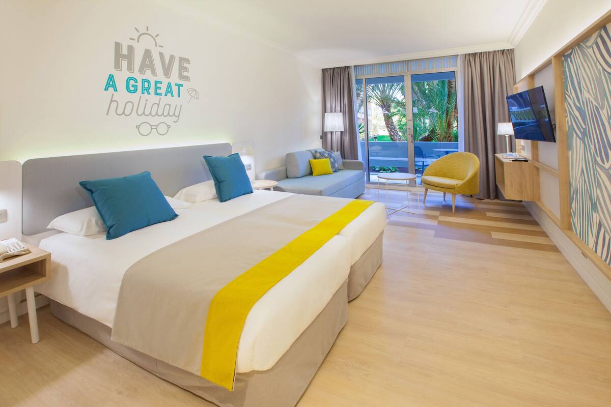 Habitación de hotel con una cama grande, un sofá, una silla, un televisor y un balcón con cortinas.