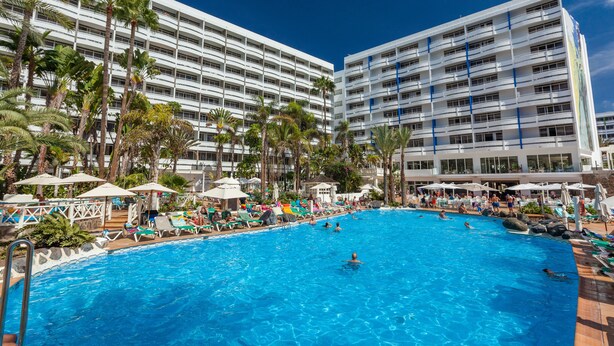 A large swimming pool surrounded by palm trees and lounge chairs, with a multi-story hotel building in the background.