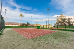 Cancha de tenis al aire libre con red, rodeada por una cerca y palmeras al fondo.