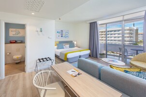 Ett modernt hotellrum med en säng, en soffa och en matplats. På väggen hänger två skyltar, en som uppmanar till vattenbesparing och en annan som handlar om kärlek. Rummet har en balkong med utsikt över staden.