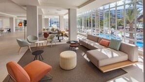 A spacious lobby with a large window overlooking a pool area, modern seating arrangements, and a central coffee table.