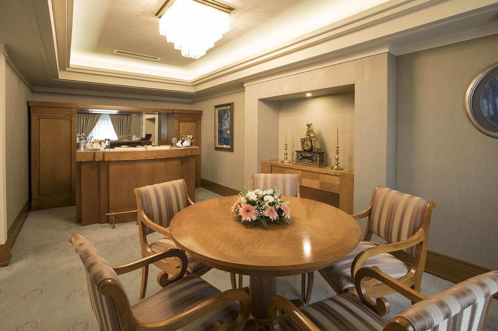 A dining area with a round wooden table, four upholstered chairs, a floral centerpiece, a wooden bar with a mirror, and a framed painting on the wall.
