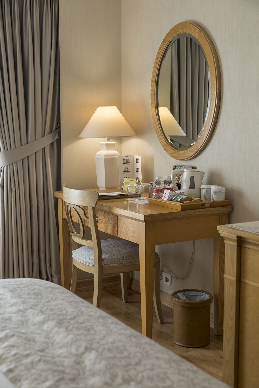 A hotel room with a wooden desk, a chair, a lamp, a mirror, and a bed.