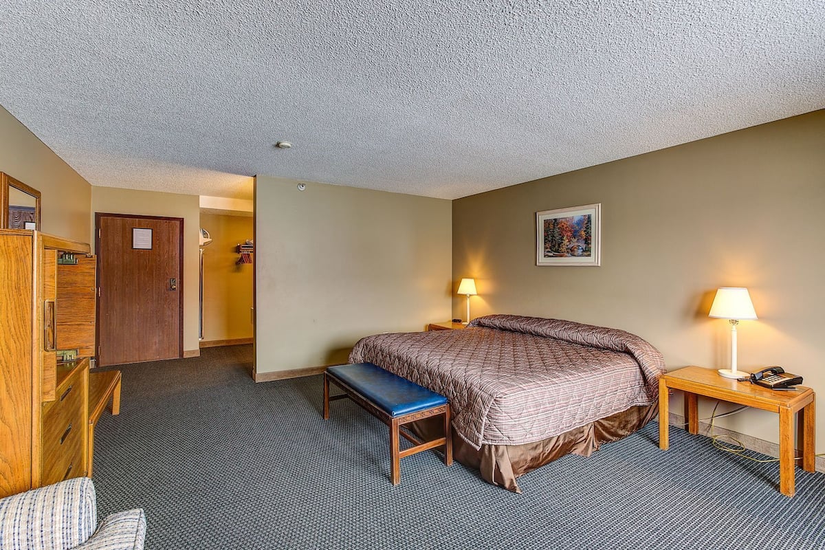 A hotel room with a bed, a desk, a chair, a bench, a door, and a painting on the wall.
