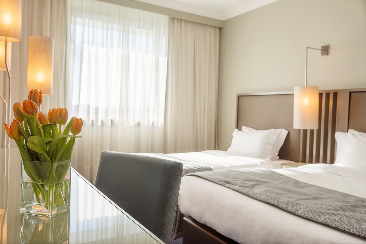 A hotel room with two beds, a glass table with a vase of flowers, and a view of the outdoors through sheer curtains.