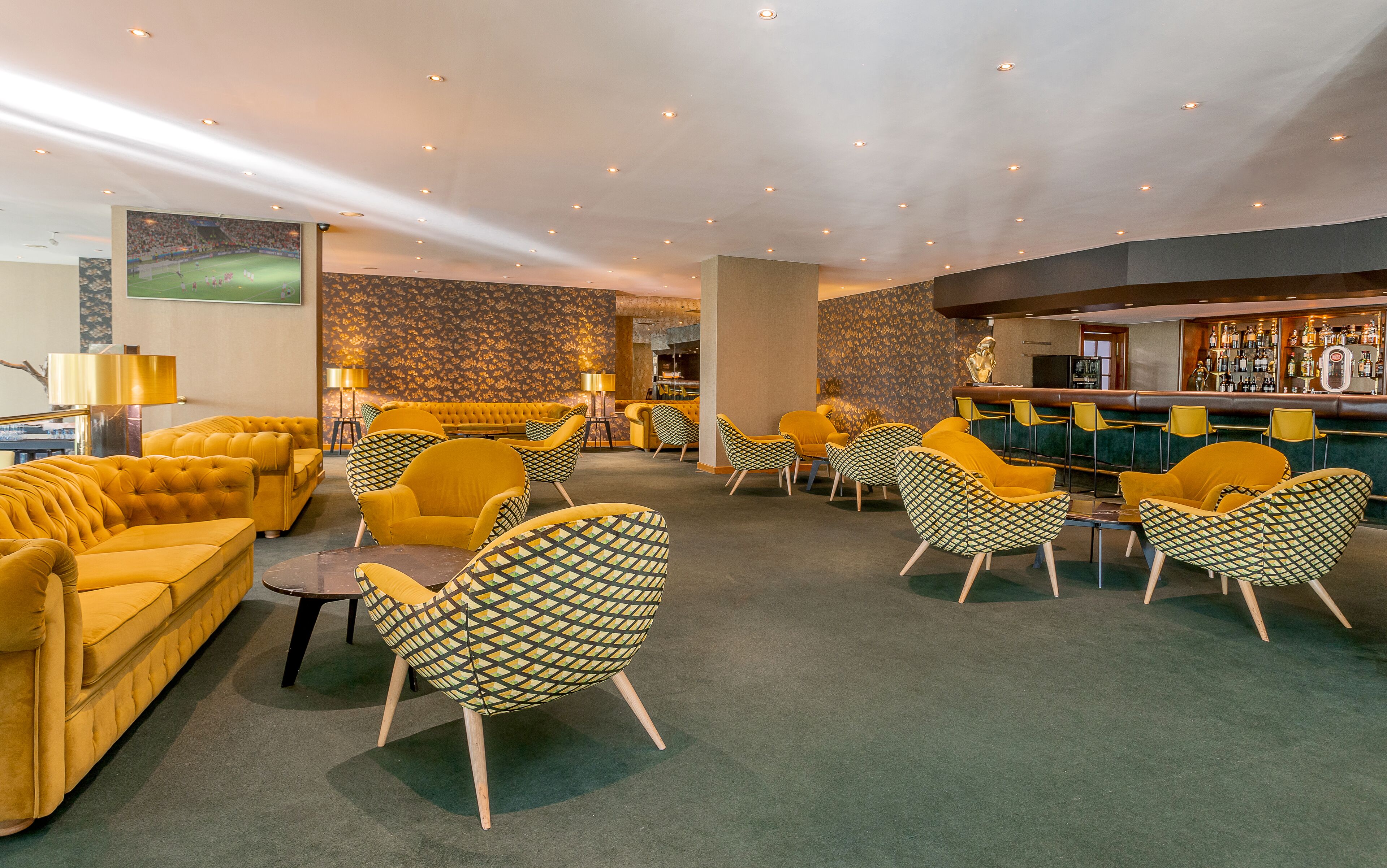 Een bar met gele stoelen en banken, een bar en een groot scherm waarop een sportevenement te zien is.