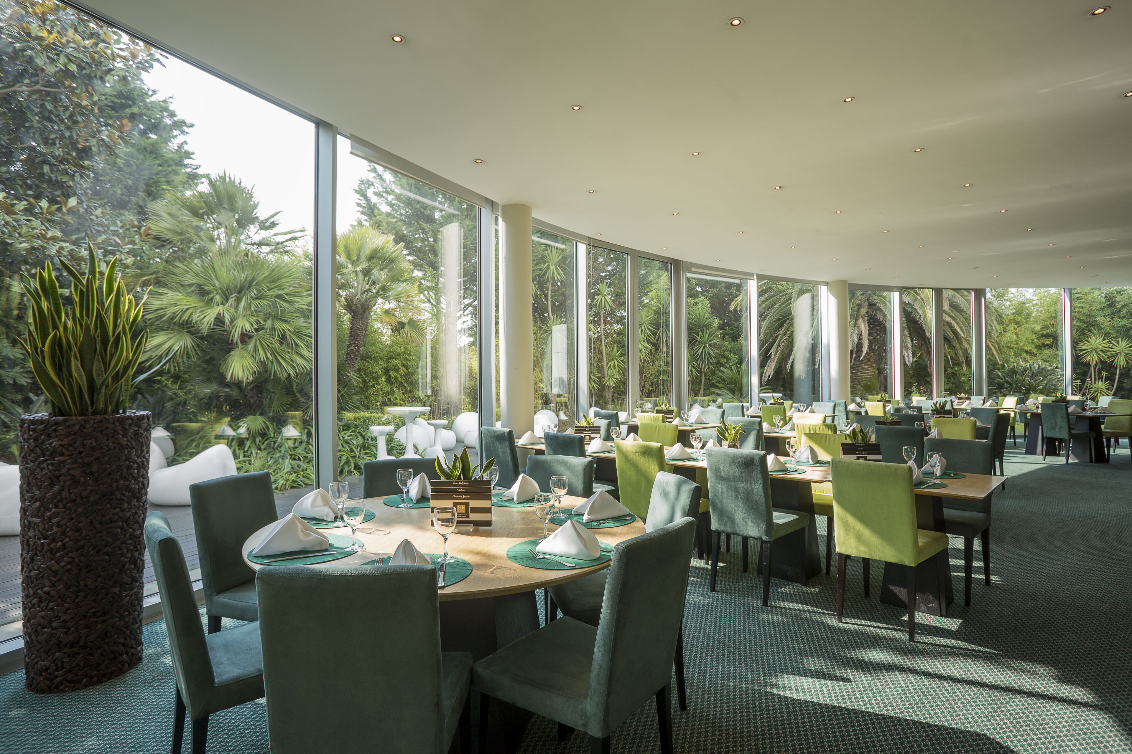 A restaurant with large windows, green chairs, and a central dining table.