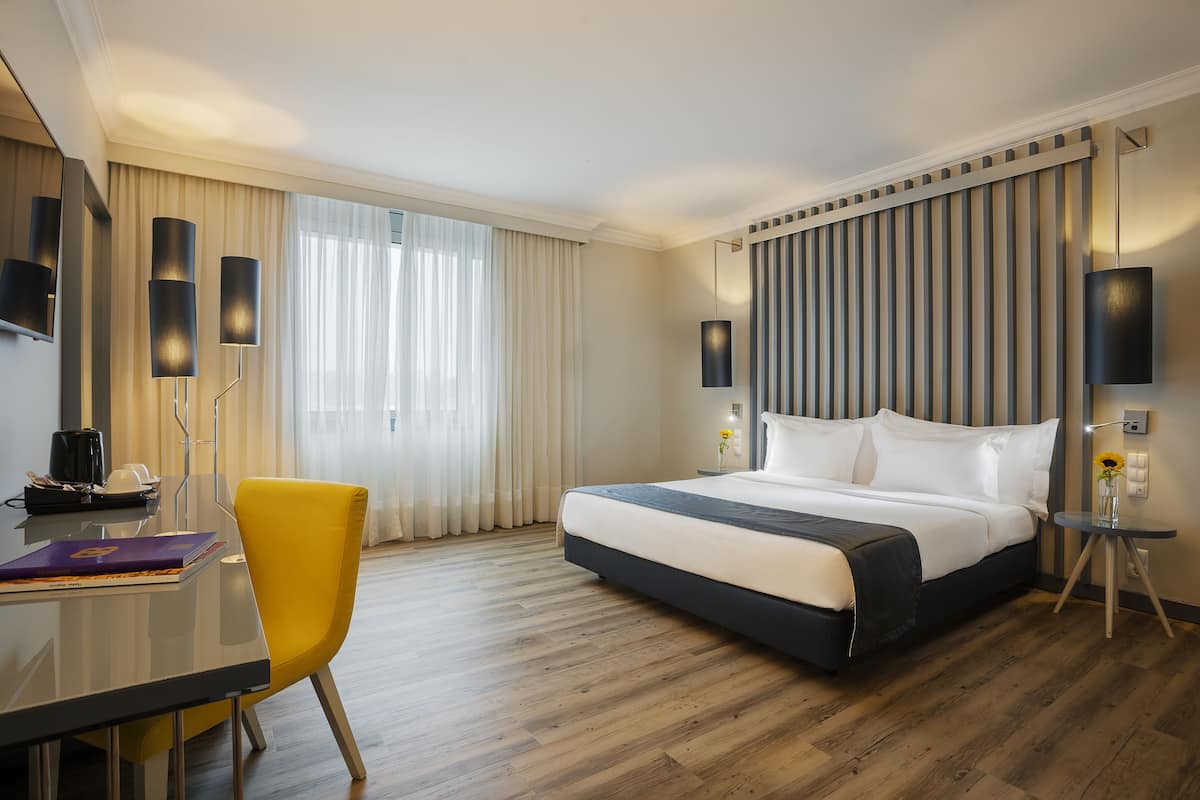 A hotel room with a large bed, a desk with a chair, a lamp, and a view of the outdoors through the window.