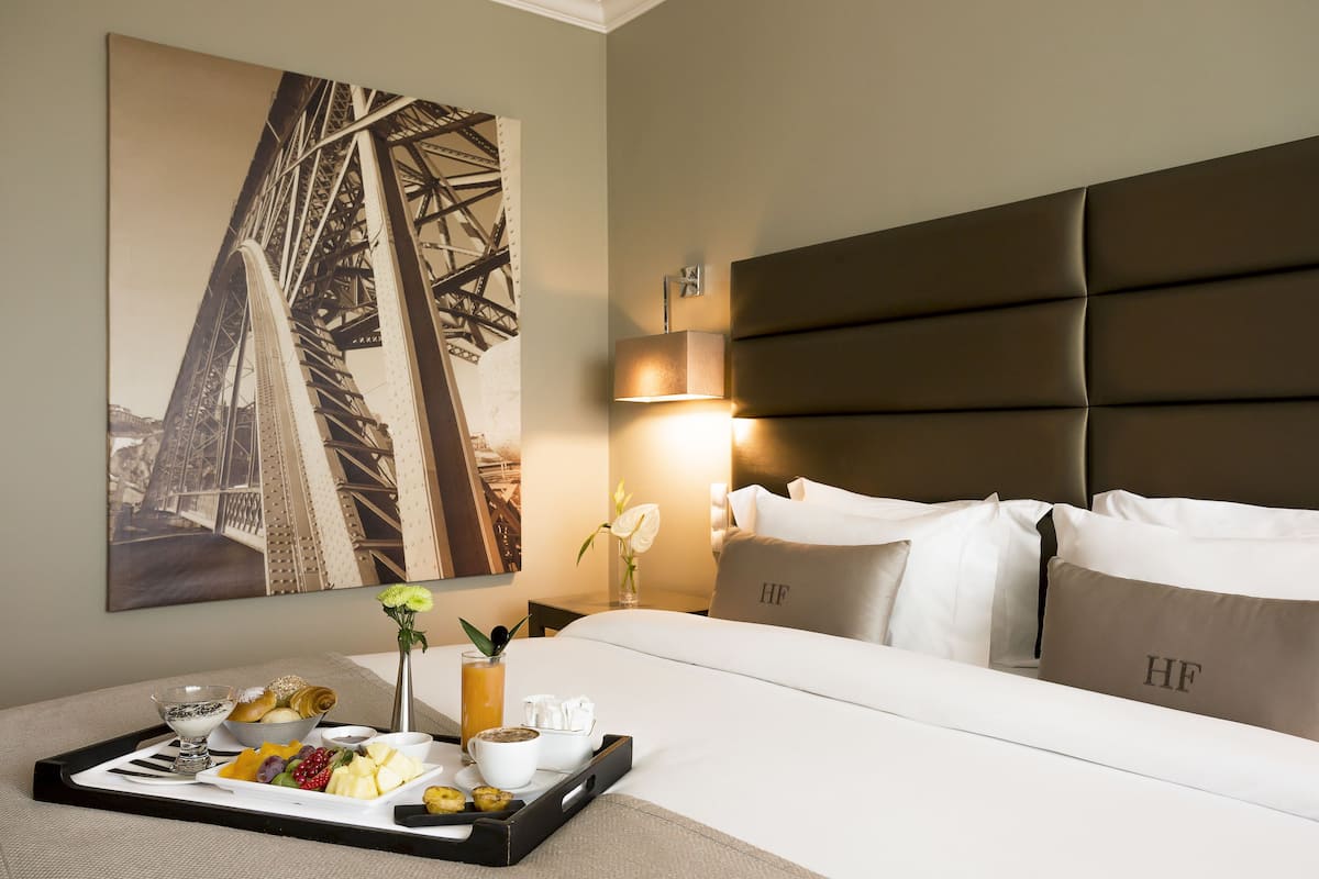 A hotel room with a bed, a tray with breakfast items, a painting of a bridge, and a nightstand with a lamp.