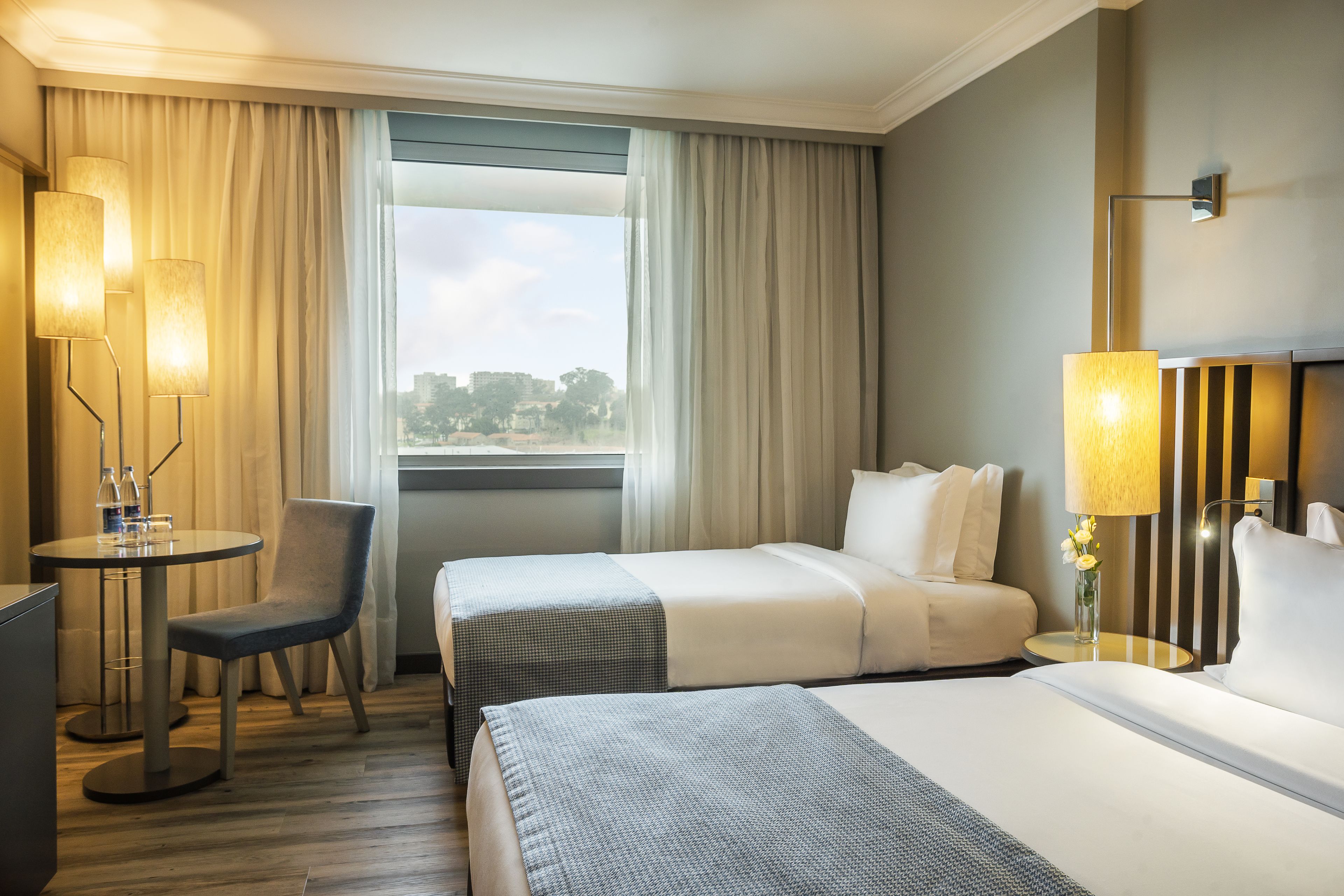 Habitación de hotel con dos camas, un escritorio con silla, una lámpara y vista a la ciudad desde la ventana.