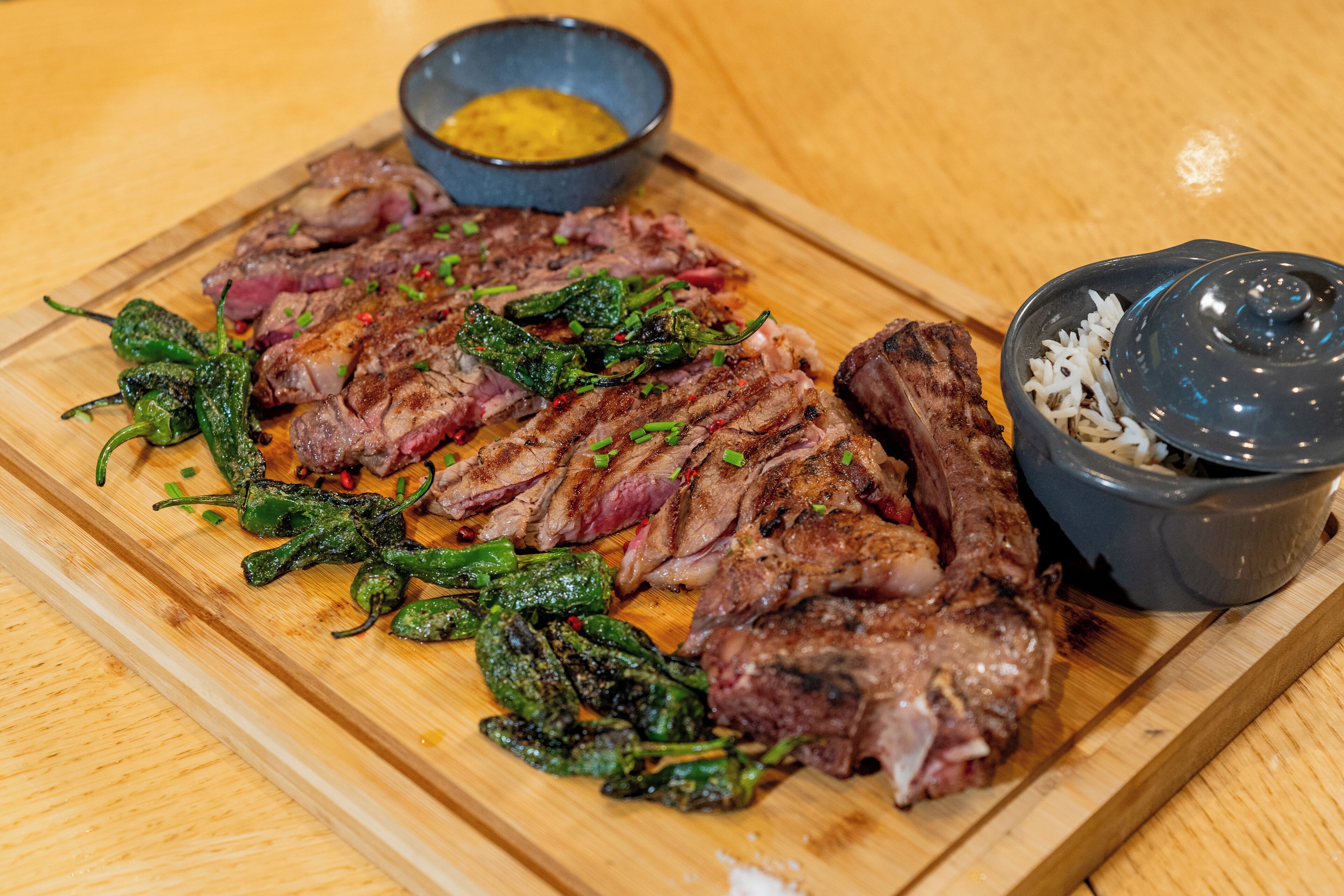 Ein Holzschnittbrett mit geschnittenem Fleisch und gegrillten Paprikaschoten, dazu eine Schüssel Soße und eine Schüssel Reis.