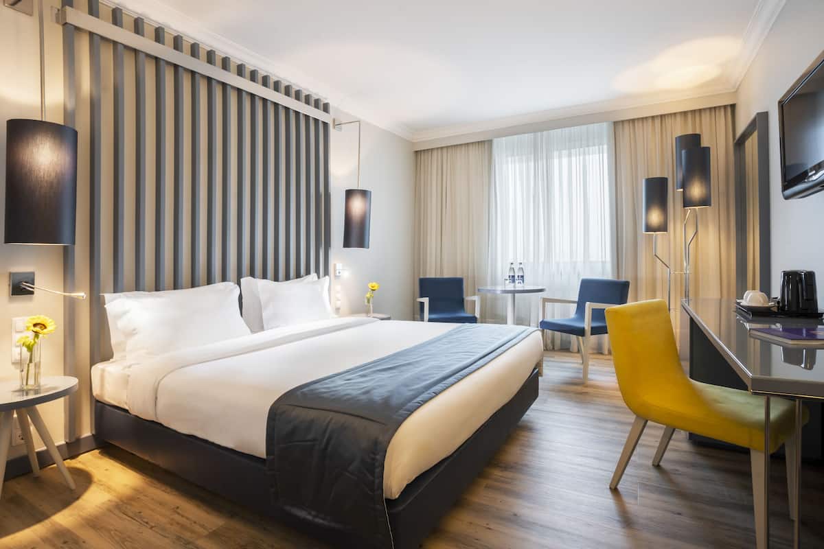 A hotel room with a large bed, a desk, a chair, a TV, and a window with curtains.