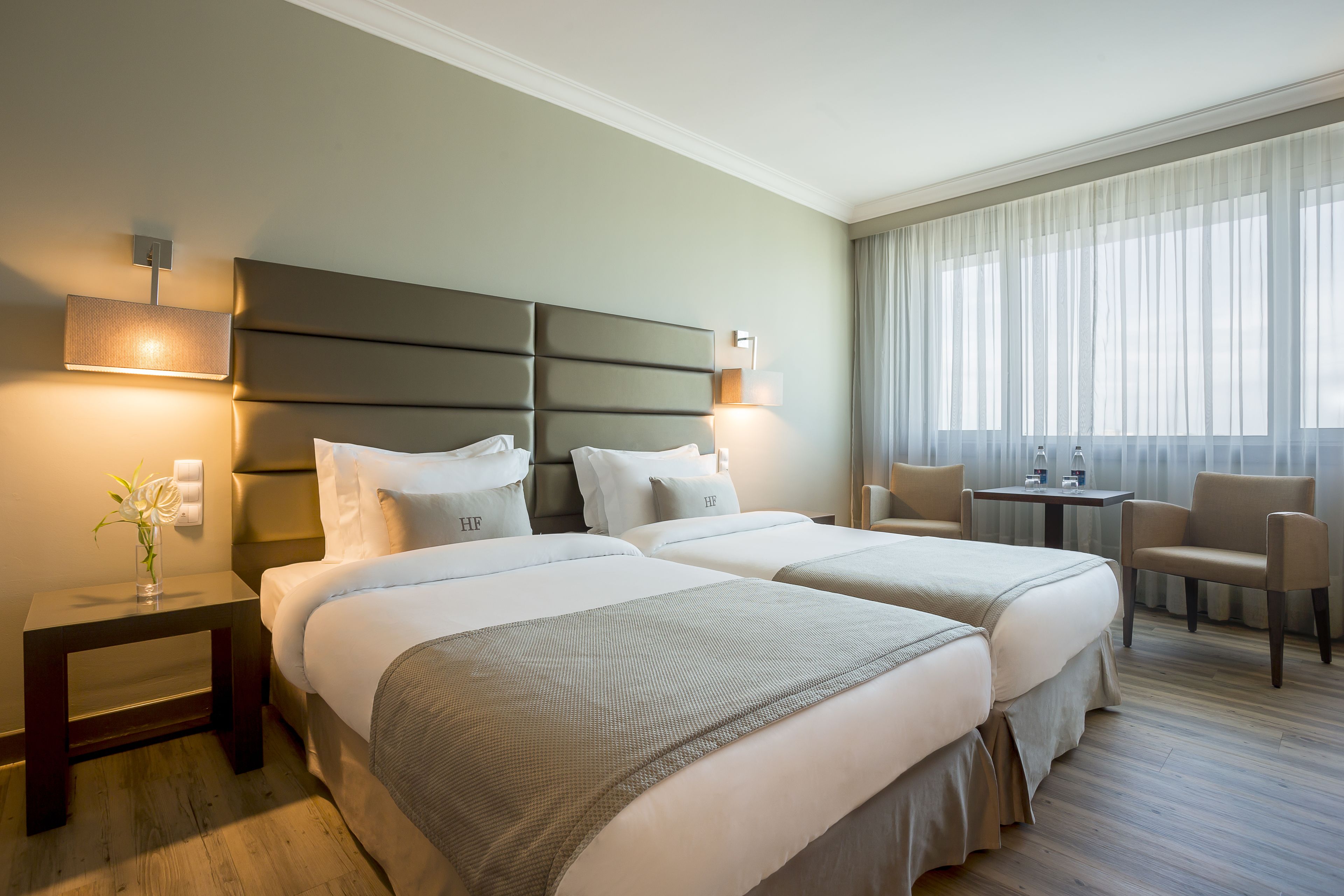 Una habitación de hotel con dos camas, una mesita pequeña, una silla y un amplio ventanal con cortinas.