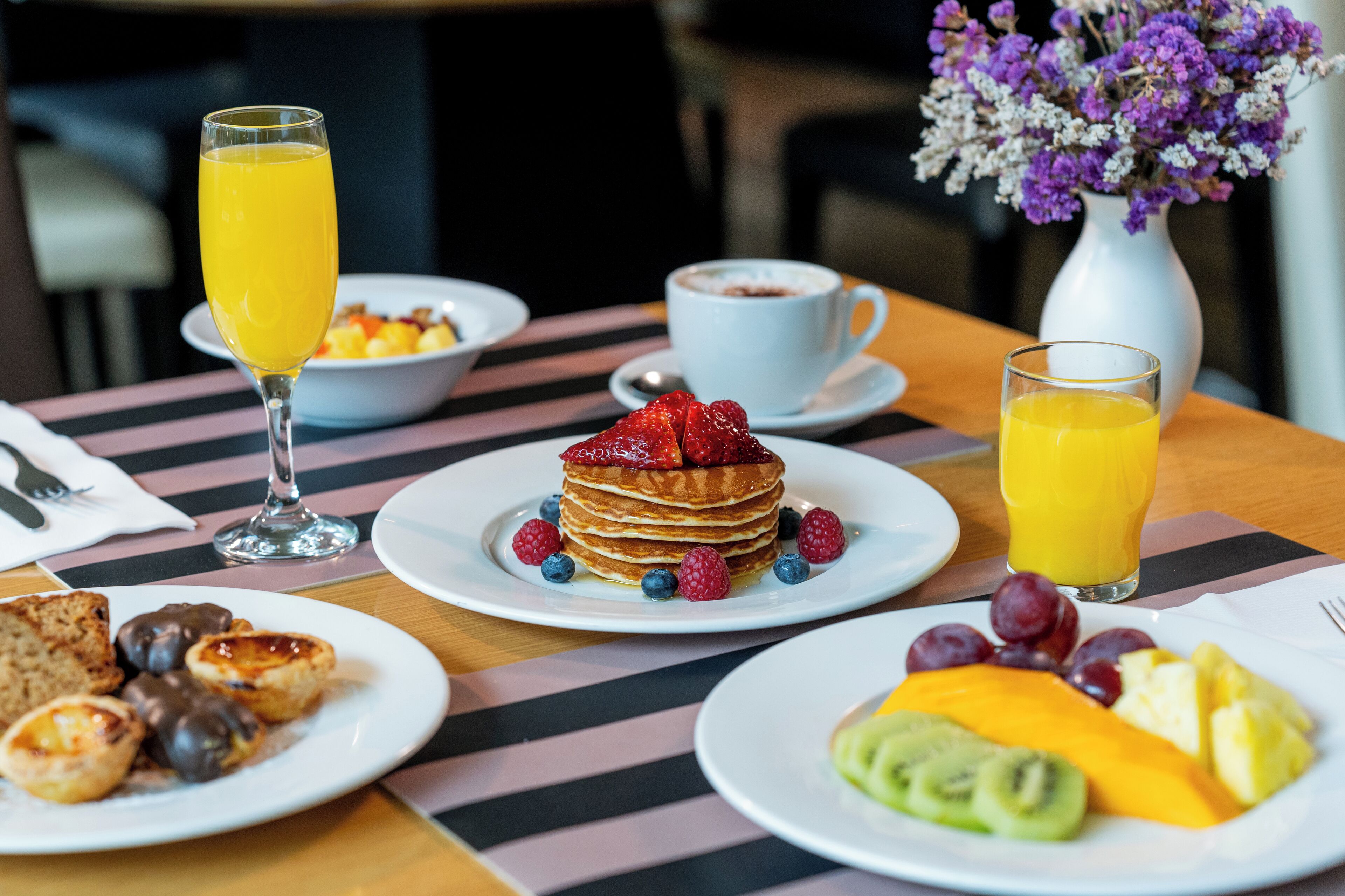 Ein Frühstückstisch mit Pfannkuchen, Obst und Getränken, bedeckt mit einer Streifenmuster-Tischdecke.