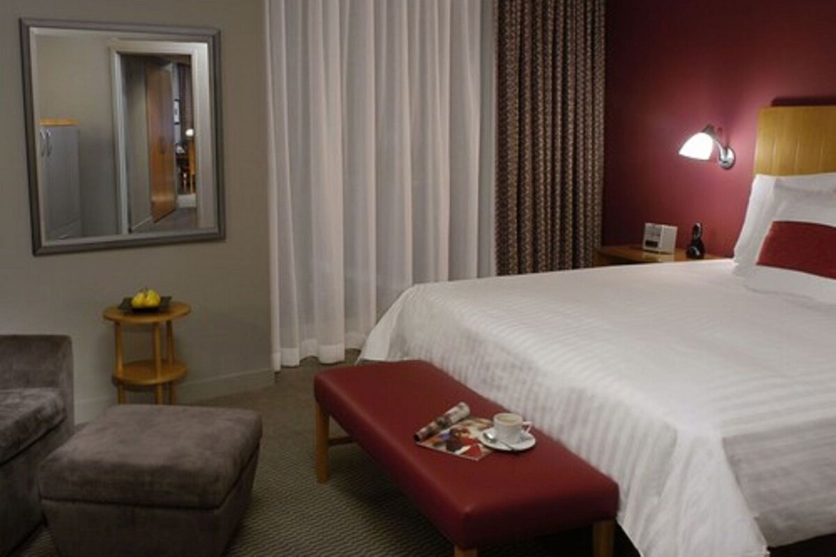 A hotel room with a bed, a bench, a small table with a fruit bowl, a mirror, and a lamp.