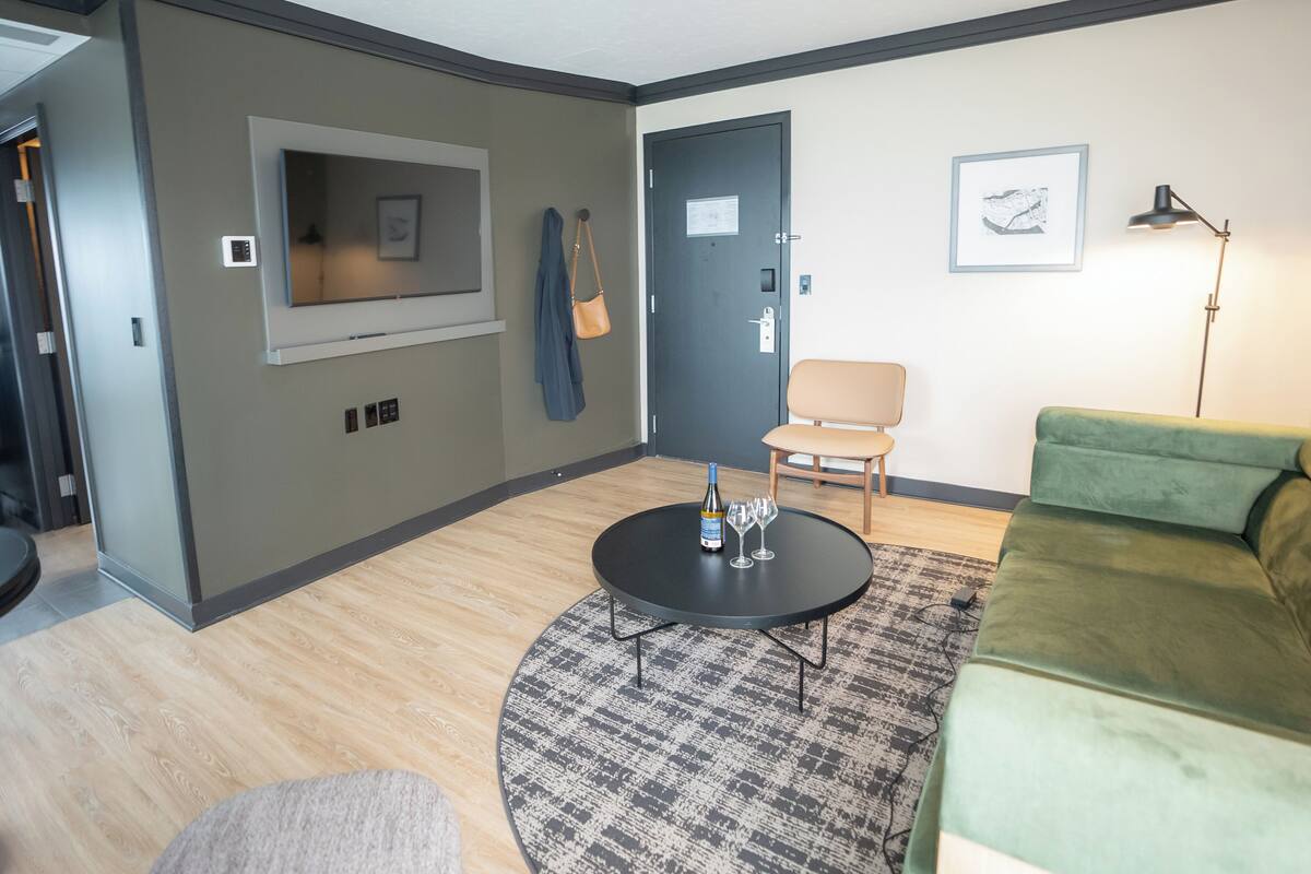 Una sala de estar moderna con un sofá verde, una mesa de centro redonda, un tapete estampado, un televisor montado en la pared, una puerta con un aviso y una silla.
