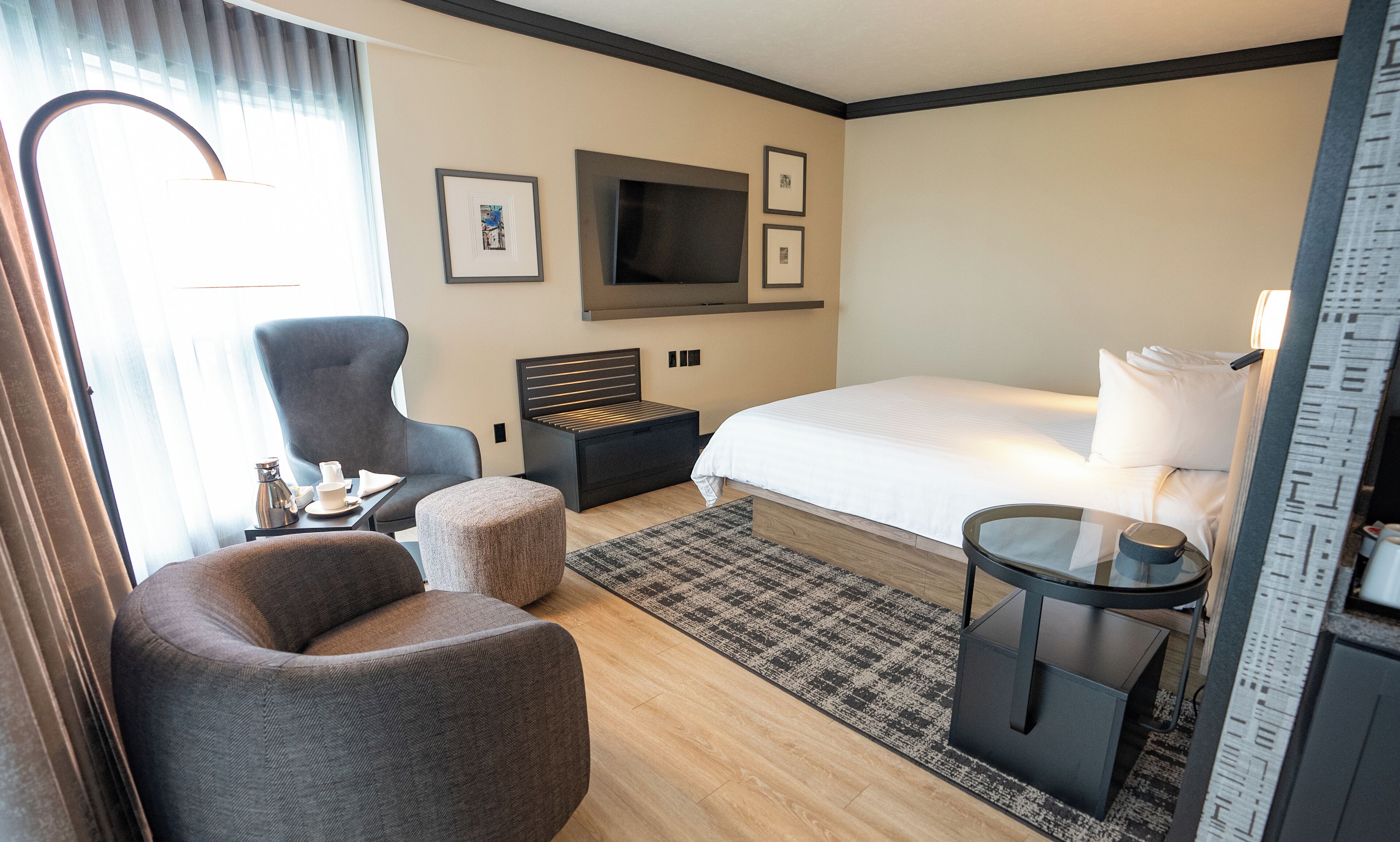 Quarto de hotel com cama, uma cadeira, uma mesinha com abajur, televisão na parede e janela com cortinas.