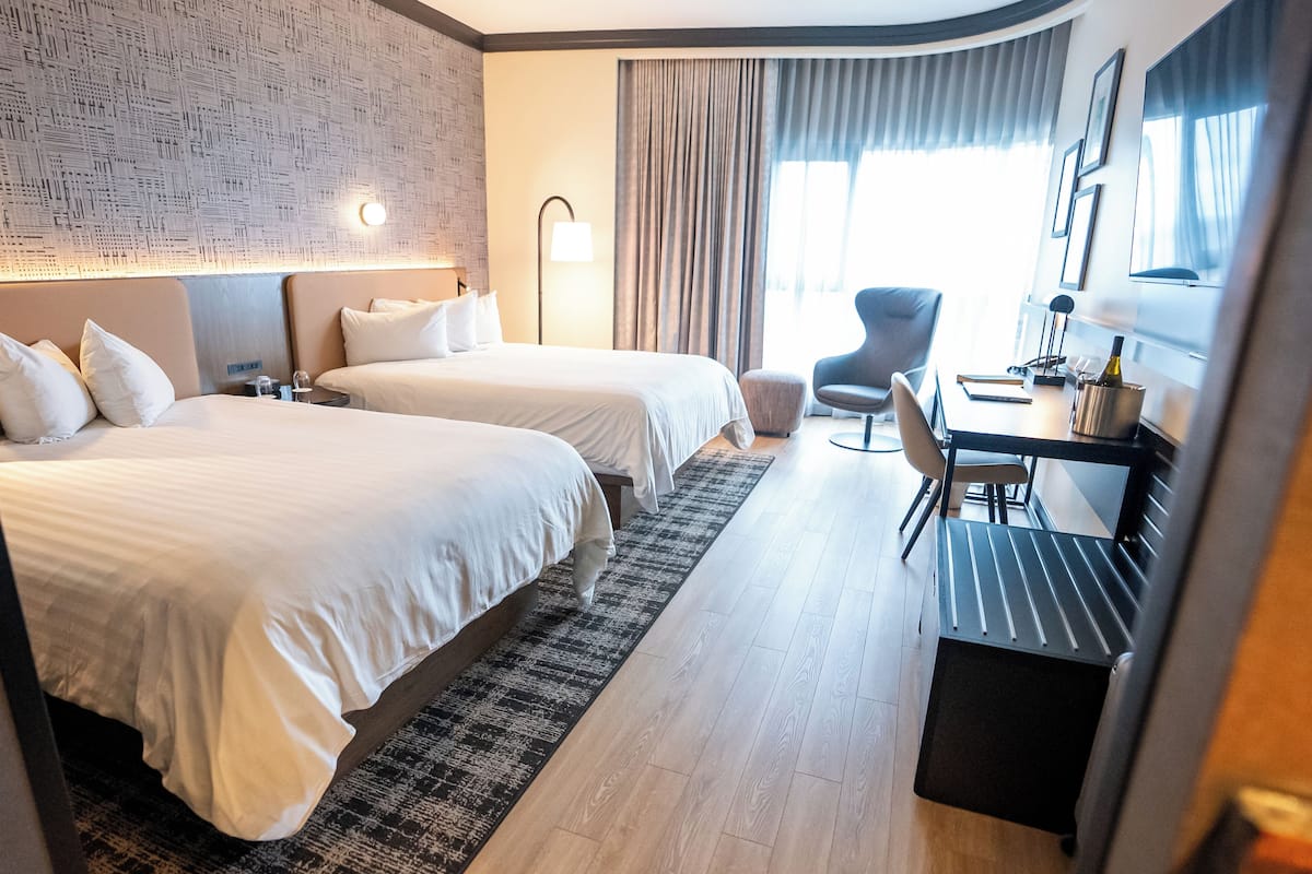 Una habitación de hotel con dos camas, un escritorio, una silla y un ventanal con cortinas.
