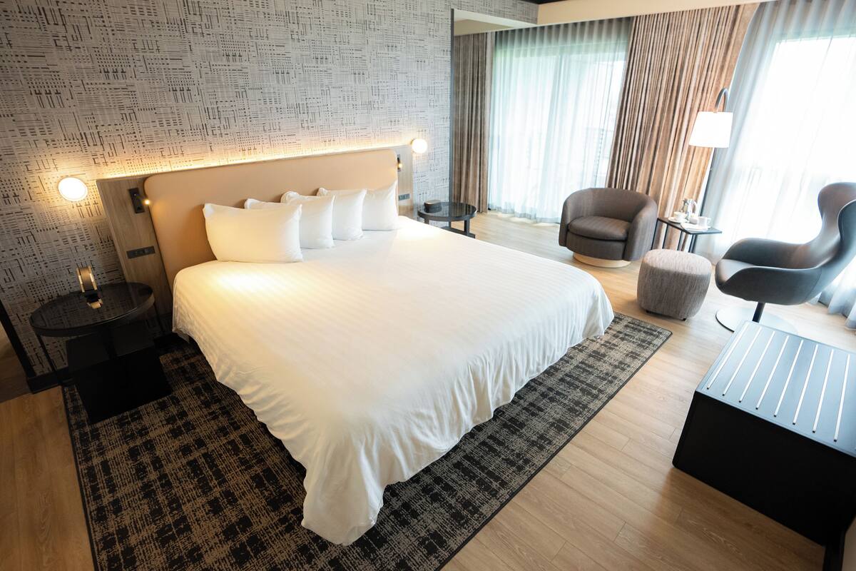 Una habitación de hotel moderna con una cama grande, una zona de estar con dos sillones, una mesita y vistas al exterior a través de amplios ventanales.