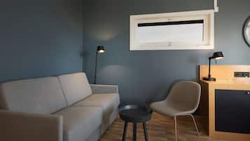 Una sala de estar moderna con un sofá gris, una mesita redonda pequeña, una silla, un escritorio con una lámpara y una ventana con persianas.