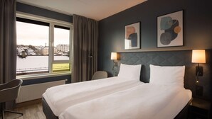 Habitación de hotel con una cama grande, una silla, una mesita de noche y vistas a la ciudad desde la ventana.