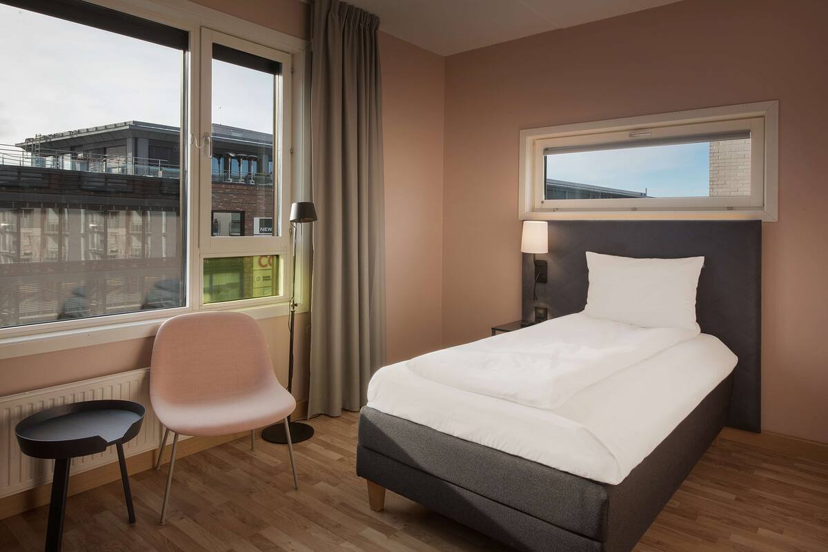 A hotel room with a bed, a chair, a lamp, and a window with a view of buildings.