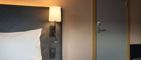 Ein Hotelzimmer mit Bett, Nachttisch mit Lampe, Bedienfeld und Tür.